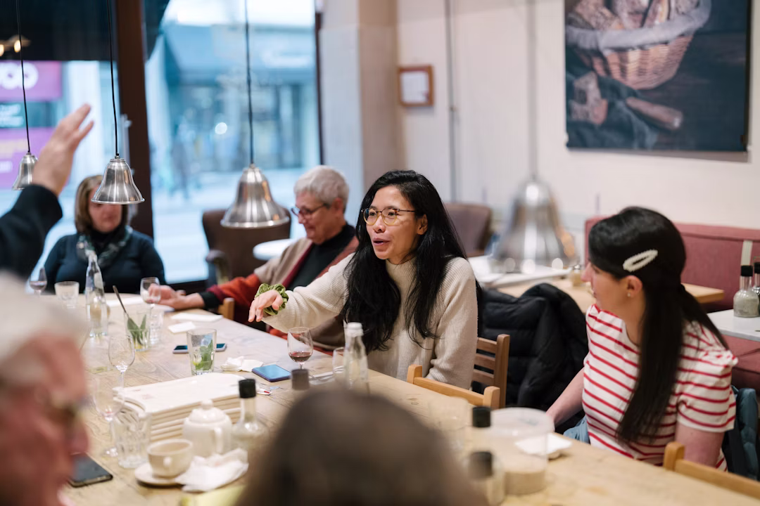 A group of people sit around a table in a cozy restaurant, talking and smiling. One woman in the center gestures while speaking, and another in a striped shirt listens beside her. Dishes, glasses, and menus are on the table.