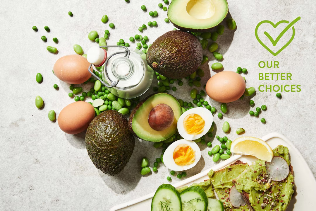 Avocados, eggs (whole and halved), green peas, and a glass bottle of milk on a light surface. A plate with avocado toast, cucumbers, and lemon is partly visible. Text reads OUR BETTER CHOICES with a checkmark heart icon.