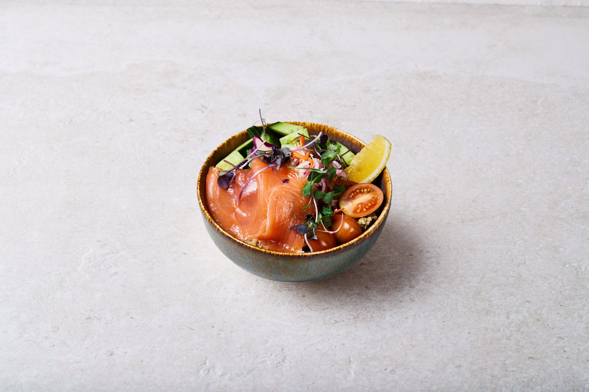 A bowl of salmon poke topped with microgreens, sliced avocado, cherry tomatoes, and a lemon wedge, served in a ceramic bowl on a light, textured surface.