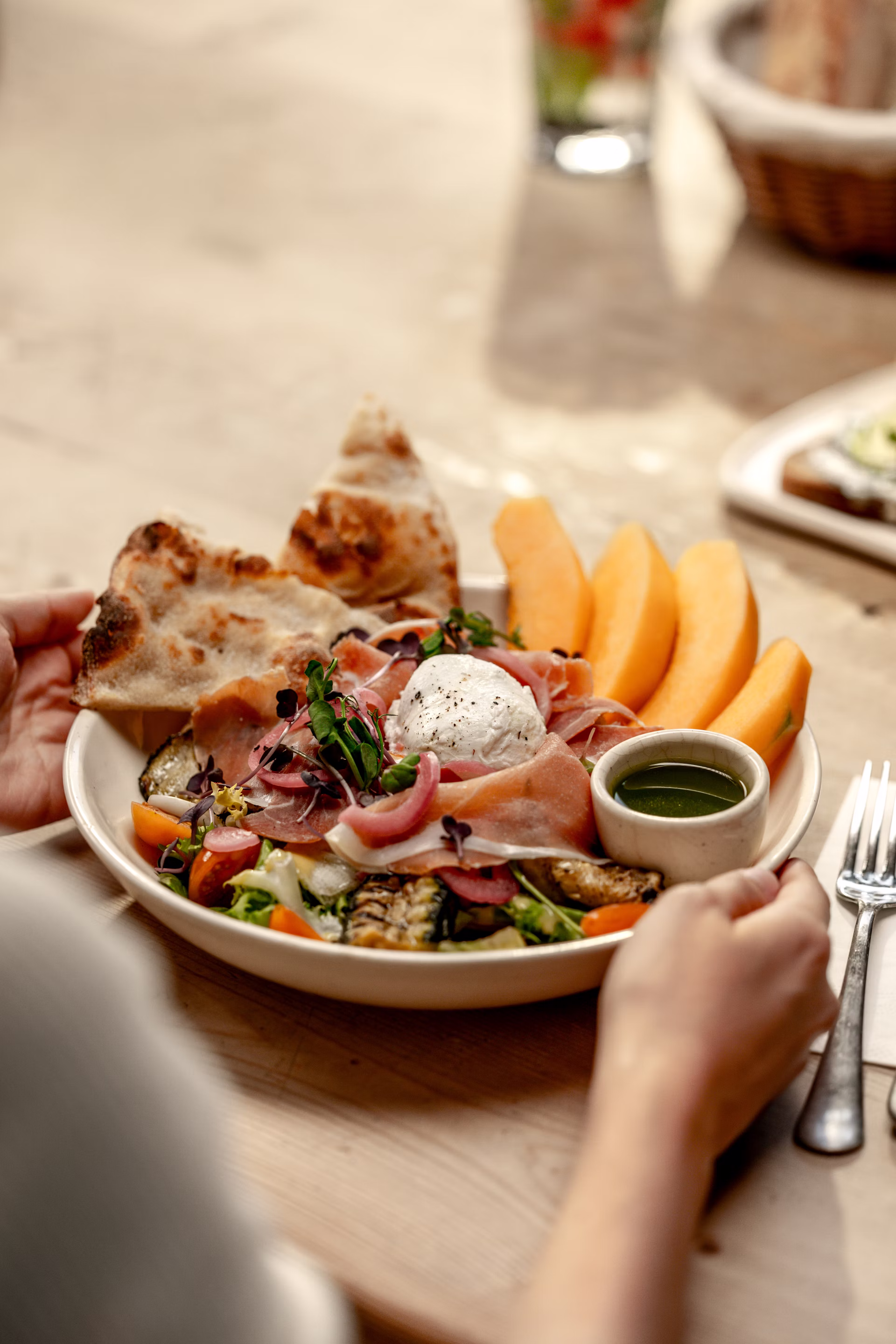 A picture of a woman holding a mediterranean salad, a plate with Charentais melon, mozzarella di bufala D.O.P., grilled zucchini, toasted flatbread, mesclun, vegan vinaigrette, pickled red onion, cherry tomatoes, sprouted cress & basil oil