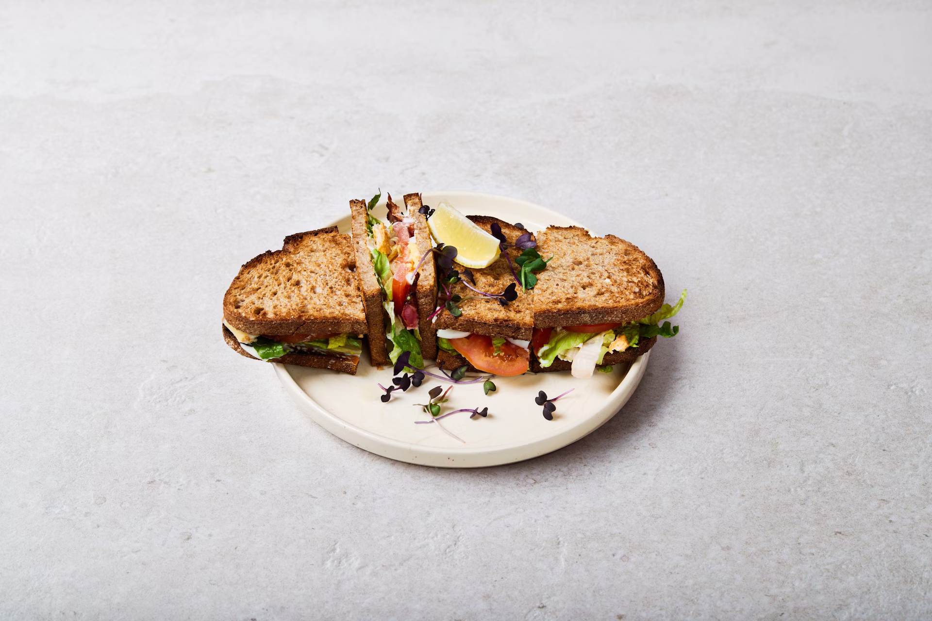 A sandwich with whole grain bread, lettuce, tomato, and other fresh ingredients is cut in half and served on a white plate, garnished with microgreens and a lemon wedge.